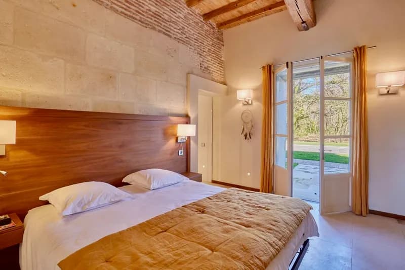 Chambre aux murs en pierre avec lit double et porte-fenêtre ouvrant sur le parc